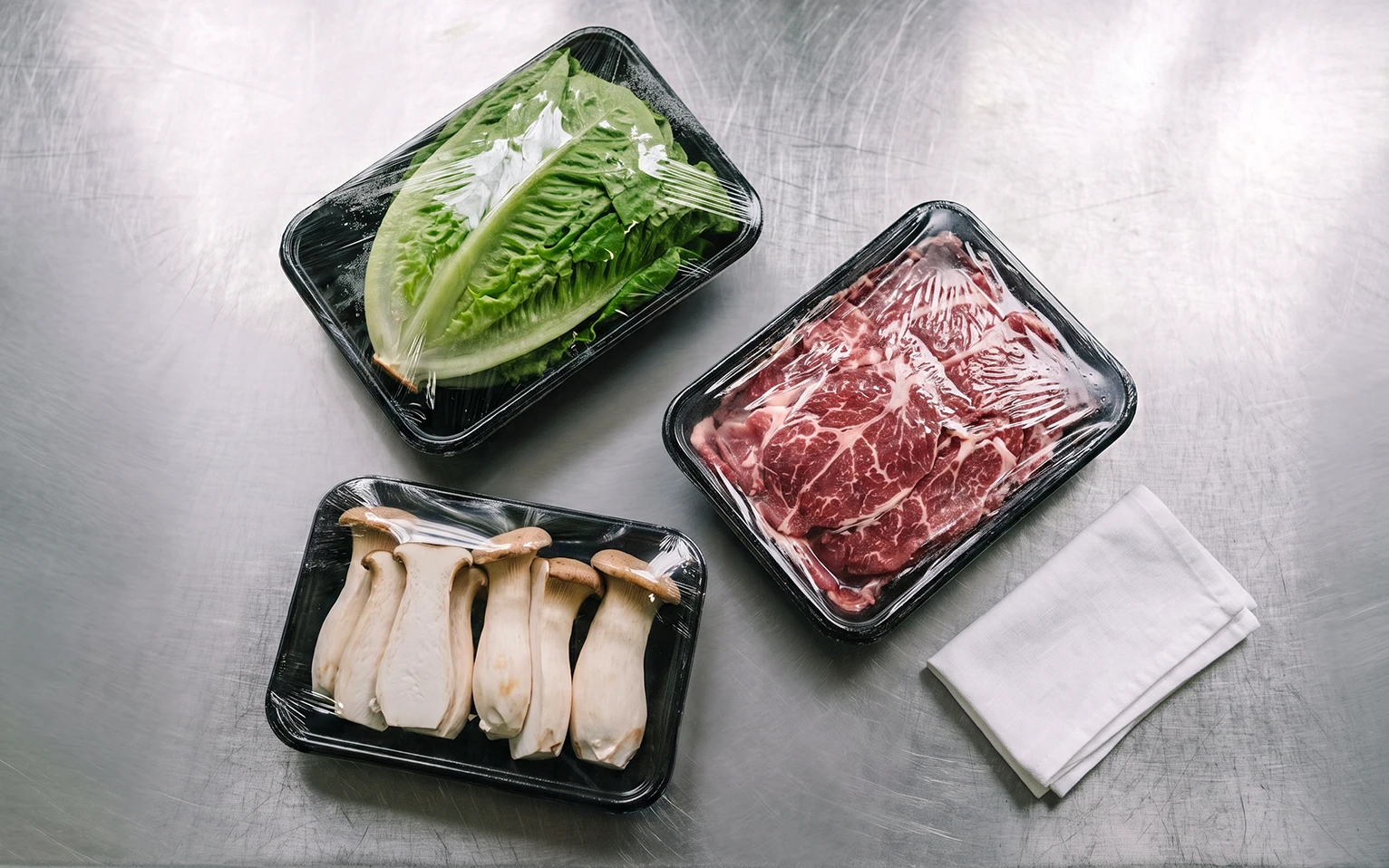Cling film tray wrapped fresh beef, lettuce, and king oyster mushrooms on a stainless steel countertop.